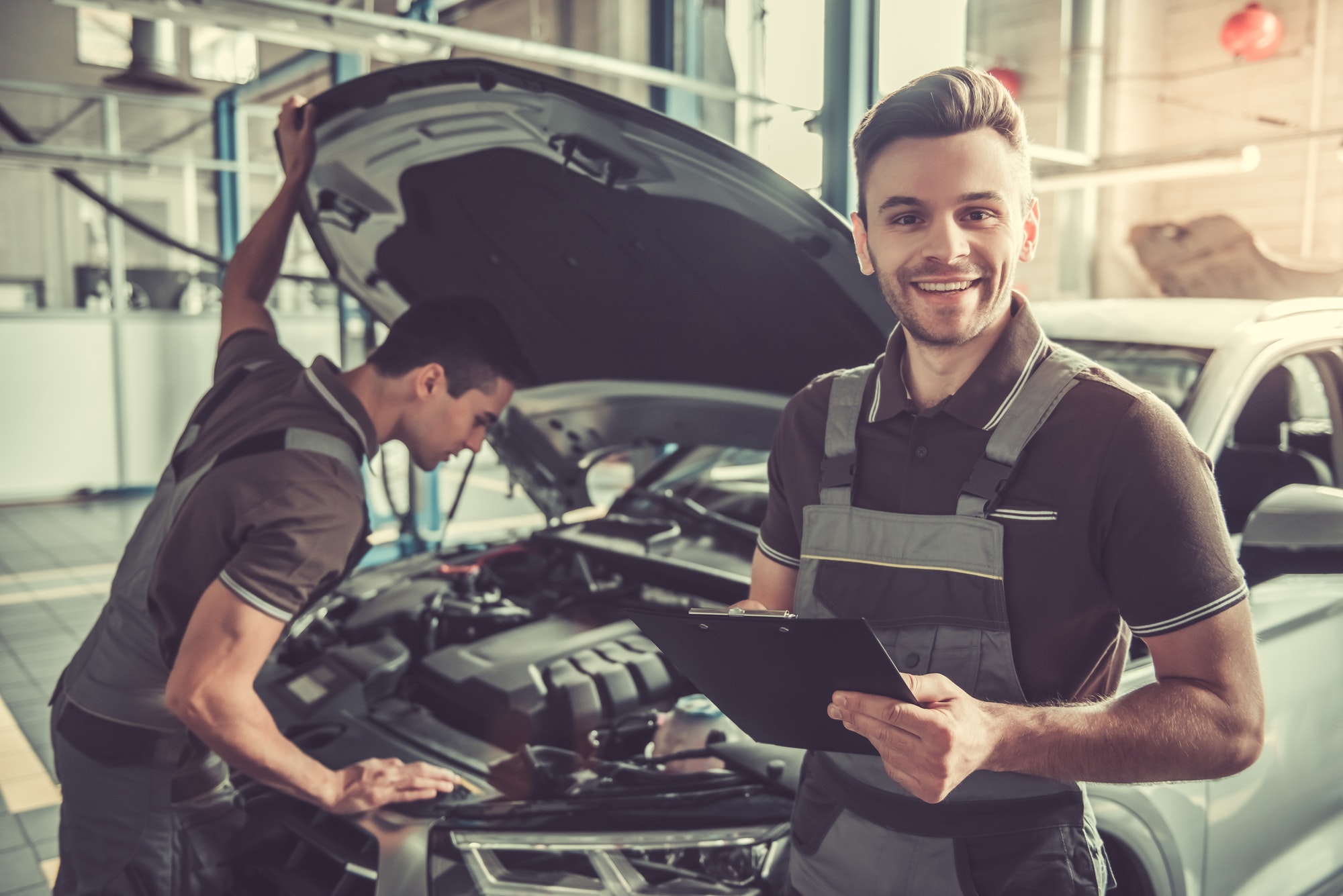 Smiling mechanic with checklist beside colleague inspecting car engine