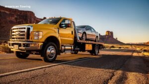 Yellow American Towing flatbed truck carrying a silver sedan on a desert highway at sunset.